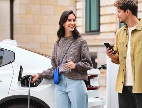 Une femme est à côté d’un véhicule électrique et regarde l’application CHARGE NOW